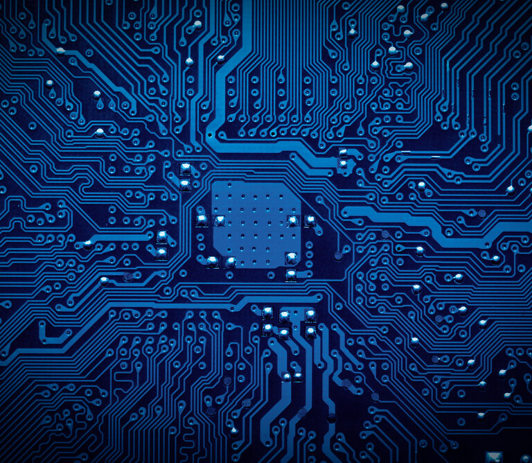 Close-up view of a blue printed circuit board showing intricate pathways and connections, with small metallic components and a central chip area, representing electronic technology.