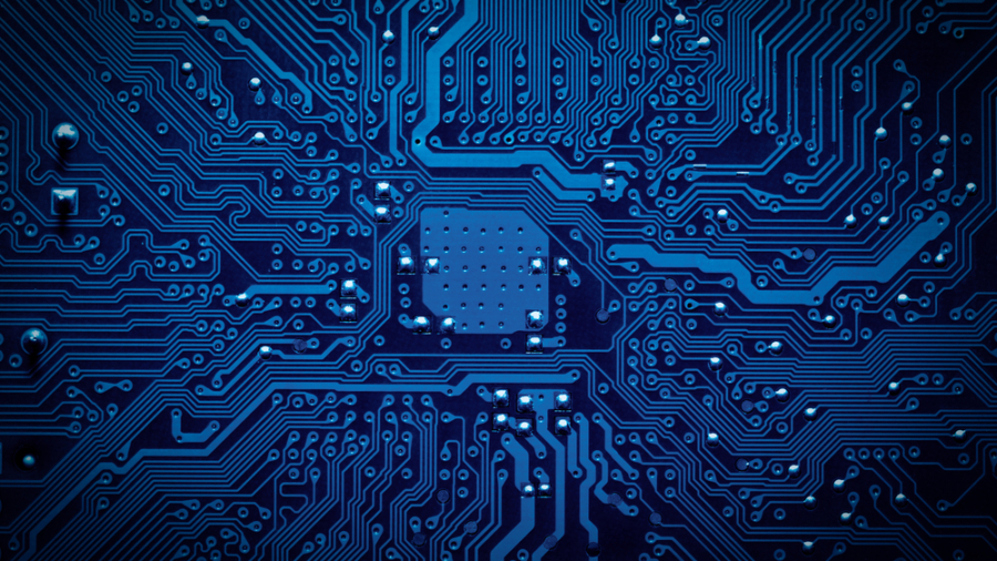 Close-up view of a blue printed circuit board showing intricate pathways and connections, with small metallic components and a central chip area, representing electronic technology.