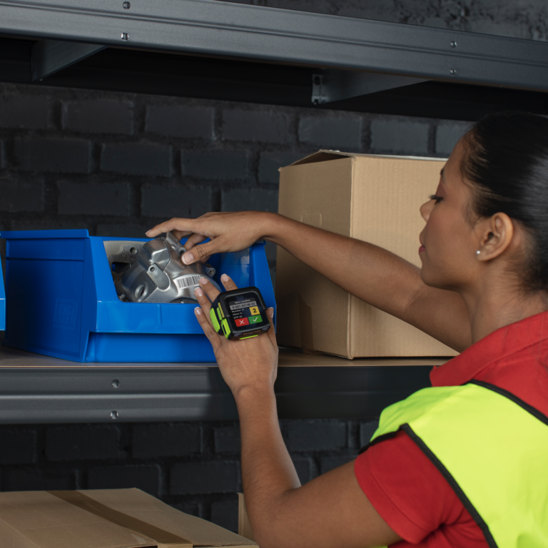 A woman wearing a high-visibility vest scans a metal part with a handheld barcode scanner while organizing blue bins and cardboard boxes on industrial shelves.