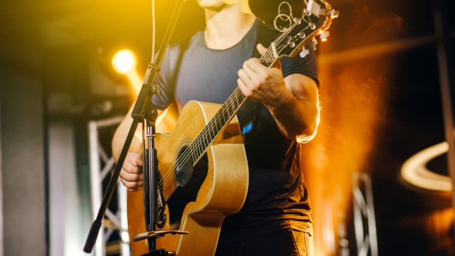 A musician in a dark t-shirt plays an acoustic guitar and sings into a microphone on a brightly lit stage with orange and yellow lights in the background.