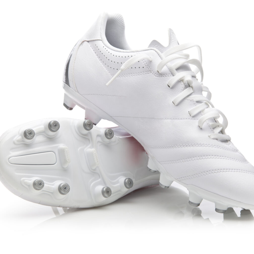 A pair of white soccer cleats, one lying on its side showing the sole with studs, and the other standing upright with laces visible, both on a white background.