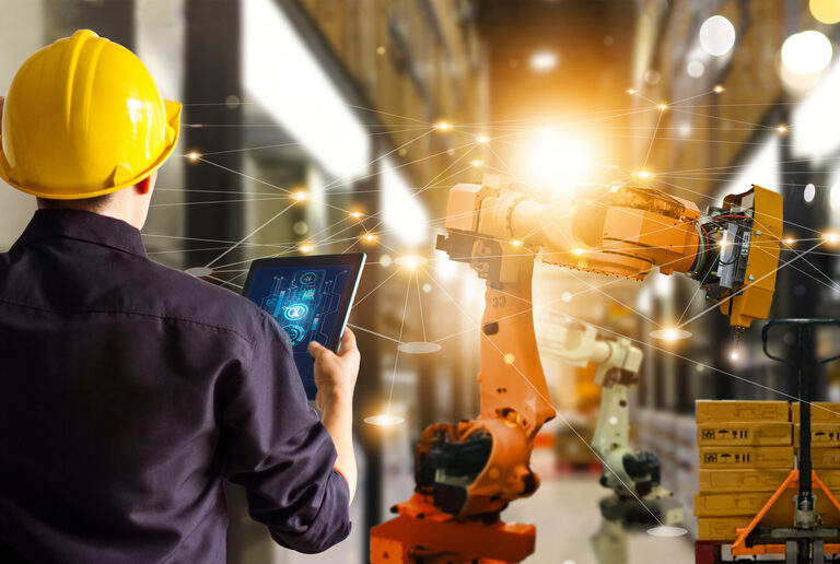 A person in a yellow hard hat uses a tablet to control a robotics arm in a warehouse, with digital network graphics overlaying the scene, symbolizing automation and smart technology.