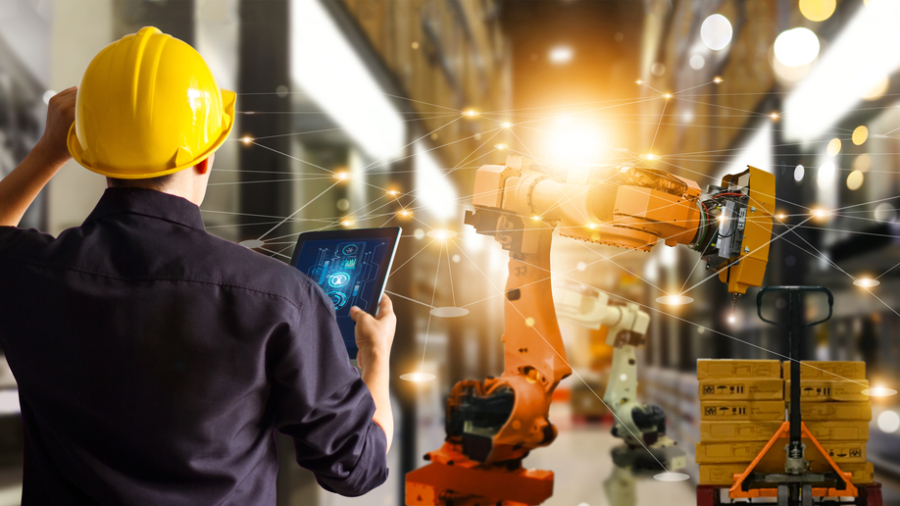 A person in a yellow hard hat uses a tablet to control a robotics arm in a warehouse, with digital network graphics overlaying the scene, symbolizing automation and smart technology.
