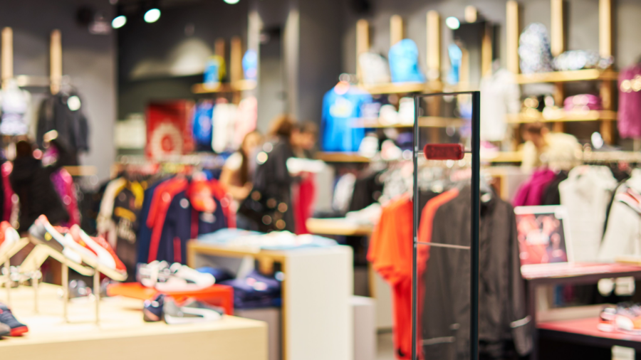 A brightly lit clothing and shoe store interior with racks of colorful clothes, shelves of sneakers, and blurred shoppers browsing in the background.