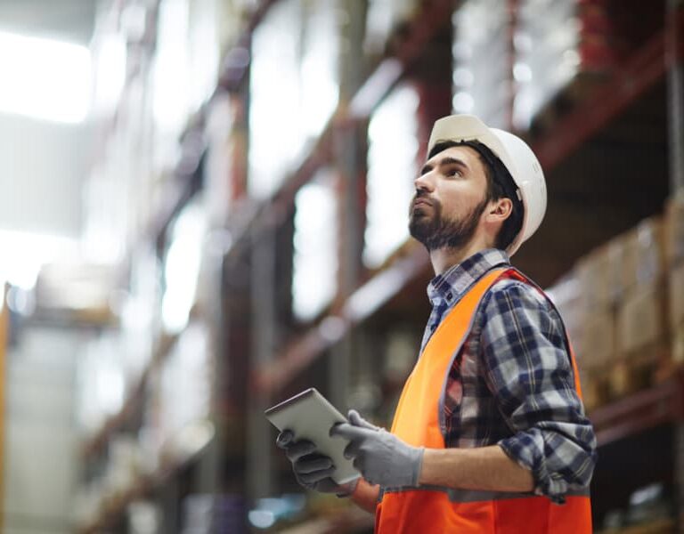 A warehouse worker wearing a safety vest and hard hat inspects shelves while holding a tablet, utilizing project management skills as he thoughtfully surveys the large warehouse filled with boxes.