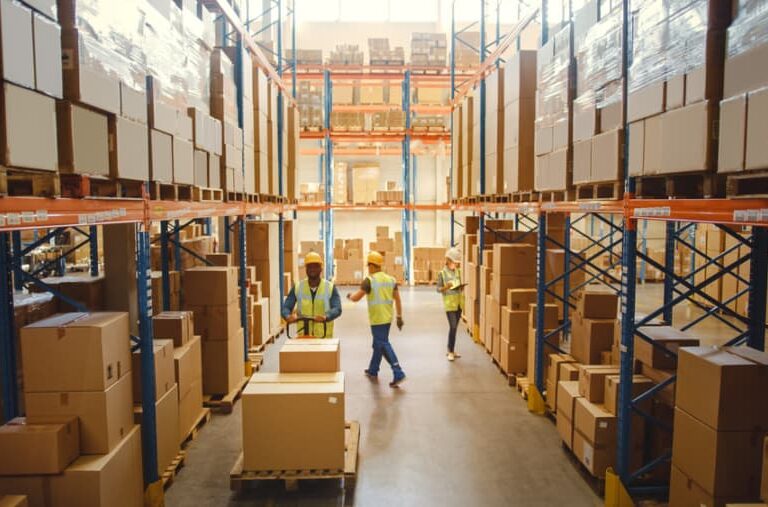 A warehouse with tall shelves filled with boxes and packages. Several workers in safety vests and helmets are organizing and moving boxes along the aisle, ensuring smooth flow as part of a Distribution Centre Operational Assessment.