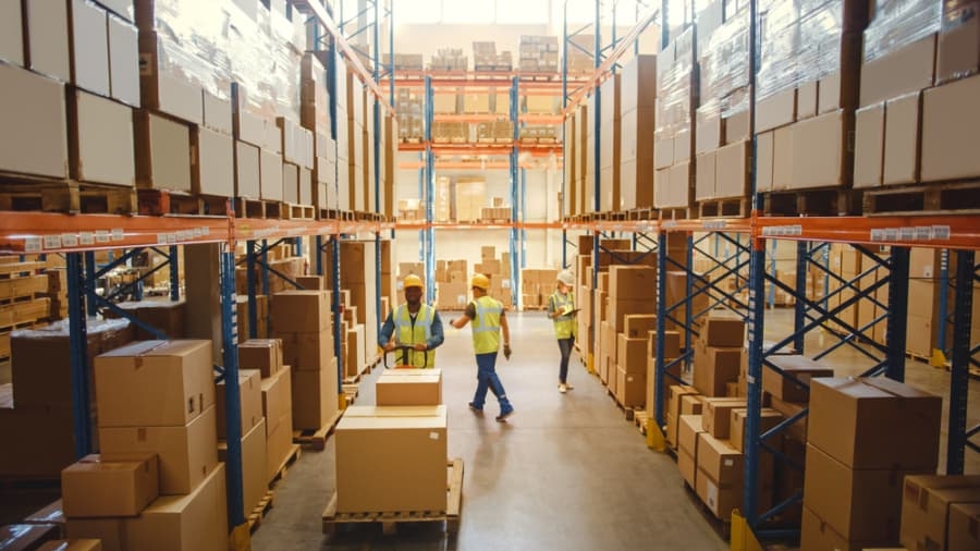 A warehouse with tall shelves filled with boxes and packages. Several workers in safety vests and helmets are organizing and moving boxes along the aisle, ensuring smooth flow as part of a Distribution Centre Operational Assessment.