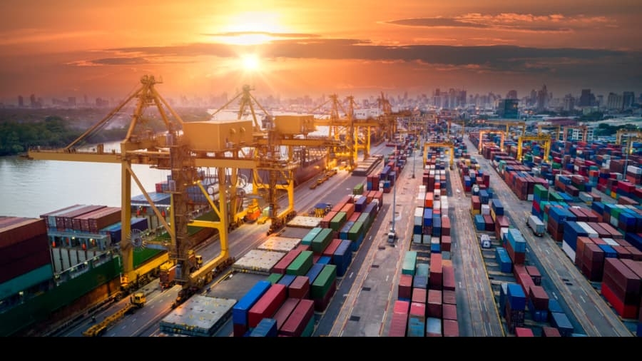 Shipping containers are stacked in rows at a busy port with large yellow cranes, as the sun sets over a city skyline in the background. The scene shows bustling industrial activity near a river.