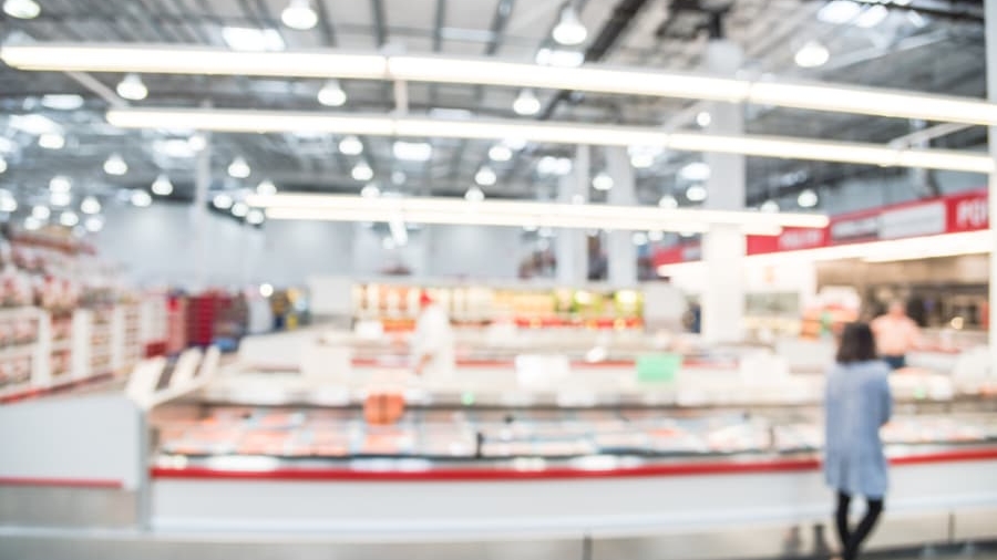 A blurry image of a person standing in a brightly lit supermarket or warehouse store, with shelves and display cases in the background. The scene is out of focus, making details difficult to distinguish.