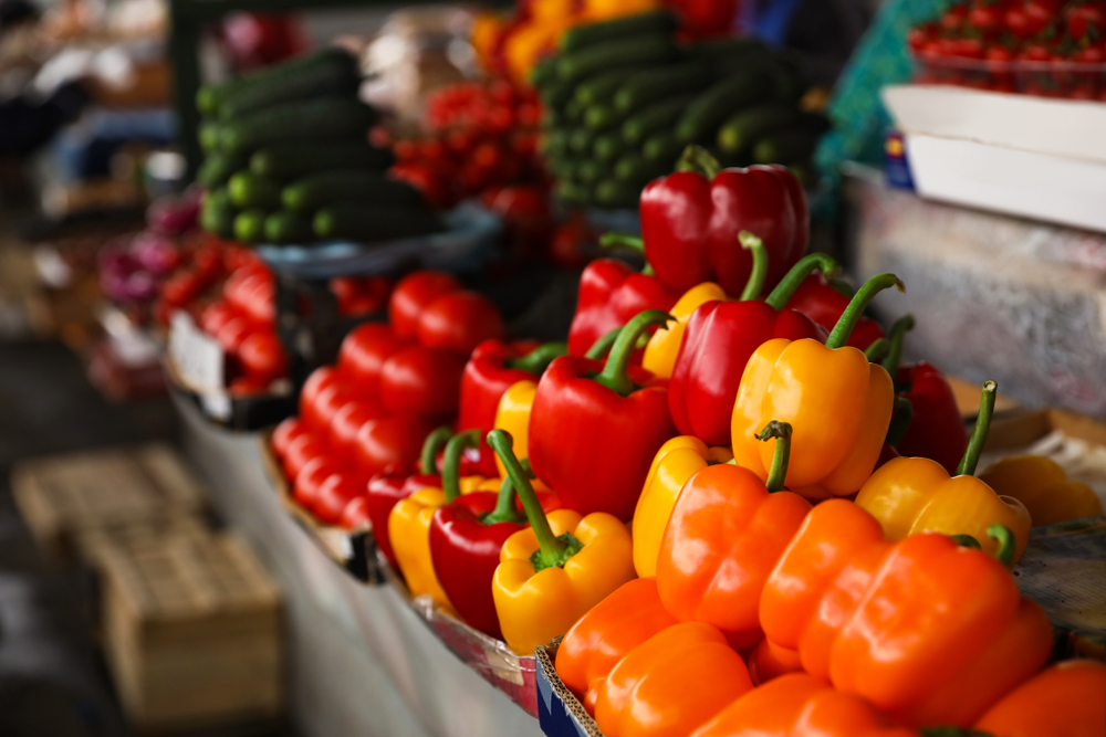Colorful bell peppers in yellow, orange, and red are stacked on a market stall, arranged as seamlessly as cloud computing organizes data. Other vegetables like cucumbers and tomatoes enhance the vibrant, fresh atmosphere.