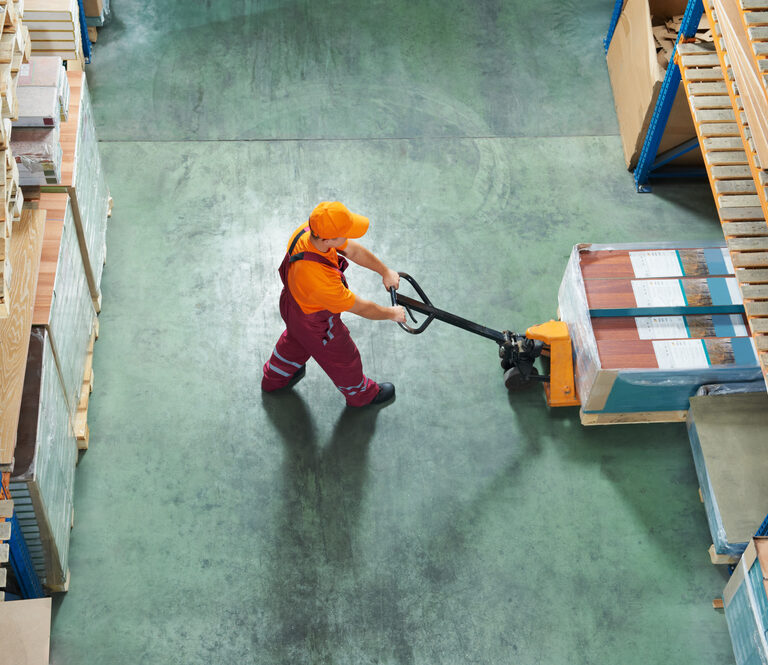 A warehouse worker in an orange shirt and cap uses a pallet jack to move a wrapped pallet between tall 3PL shelving units stocked with goods. The scene is viewed from above.