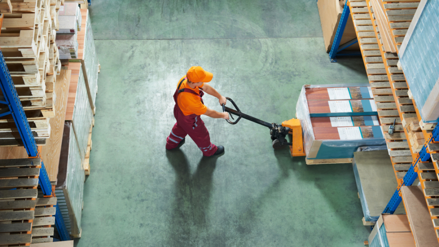 A warehouse worker in an orange shirt and cap uses a pallet jack to move a wrapped pallet between tall 3PL shelving units stocked with goods. The scene is viewed from above.