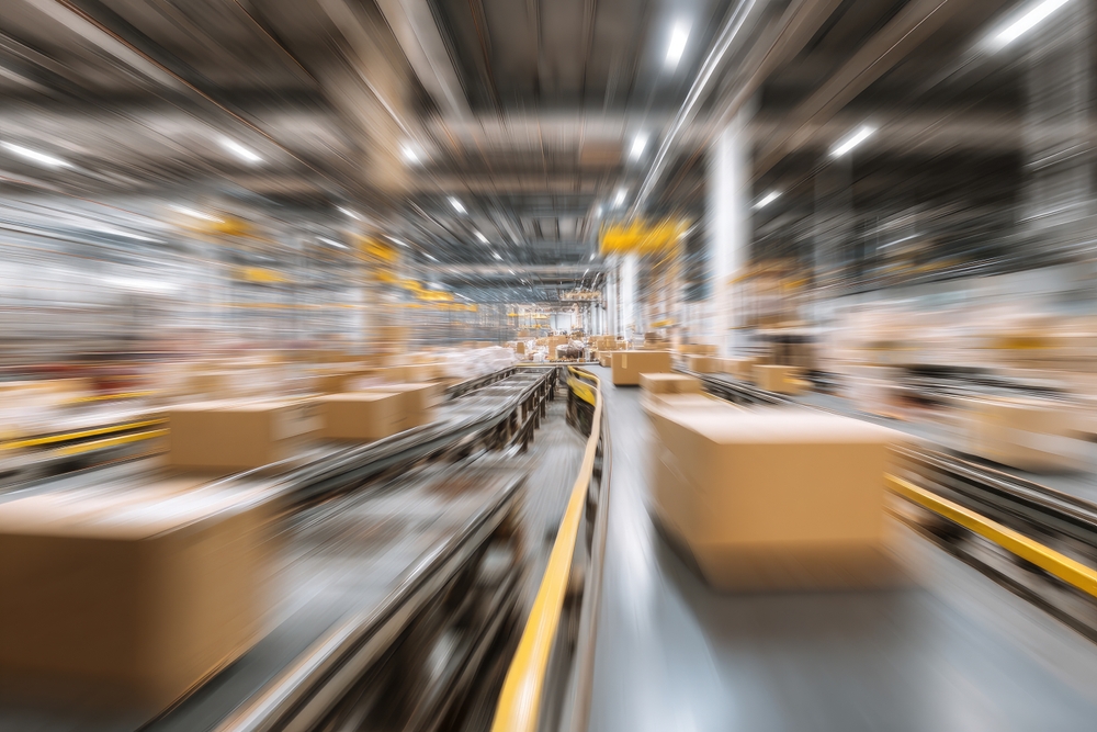Blurred image of a busy warehouse or distribution center with cardboard boxes moving quickly along conveyor belts, conveying a sense of fast-paced logistics and shipping operations.