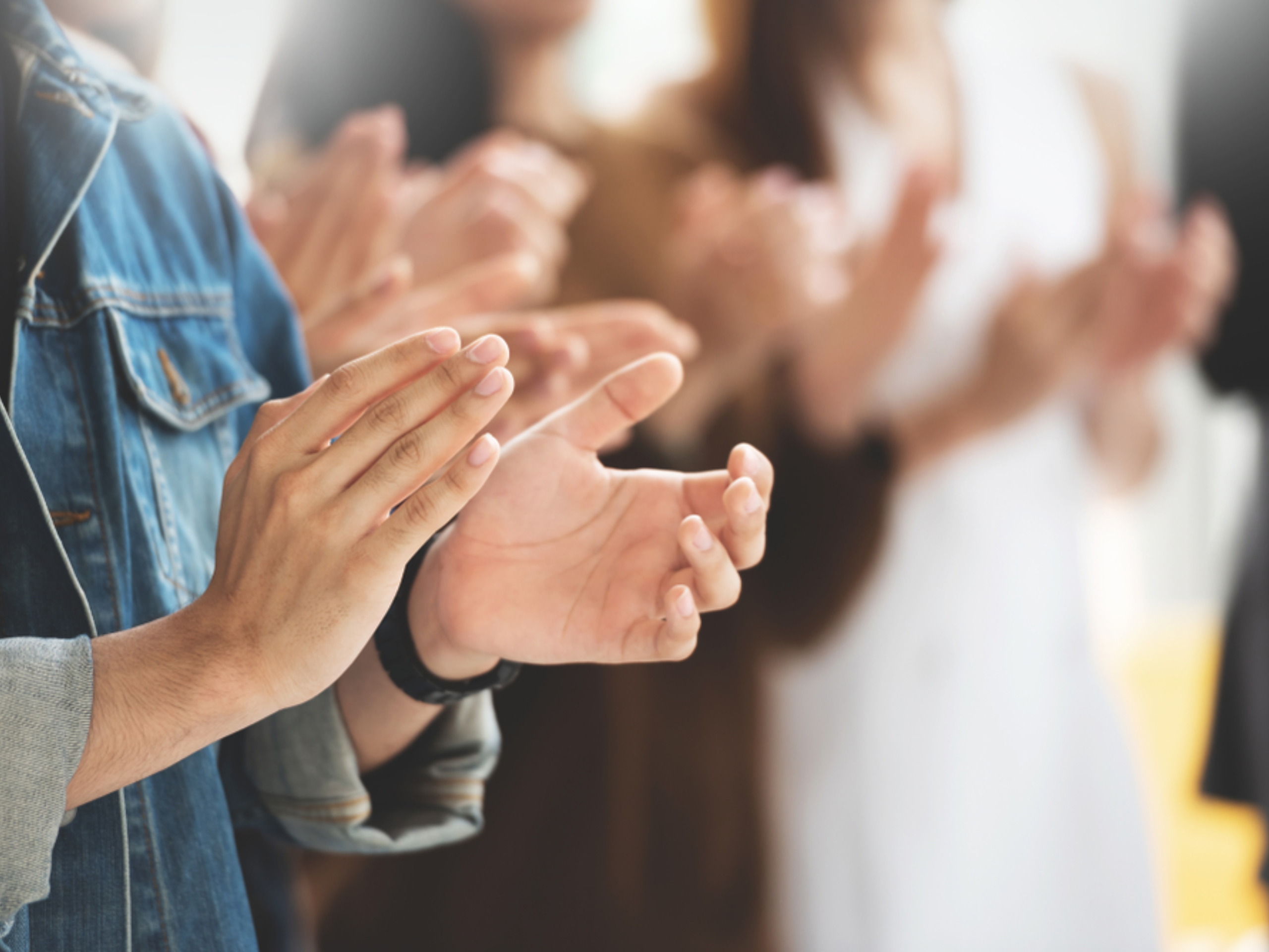 A group of people standing close together, clapping their hands in applause. The focus is on the hands and arms, with faces blurred in the background. The mood appears positive and supportive.