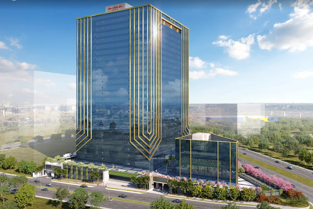 A tall, modern glass office building with gold vertical accents stands beside a tree-lined road, surrounded by greenery and a blue sky with scattered clouds. A smaller matching building sits in front, near the entrance.