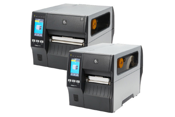 Two gray industrial label printers with digital screens and control buttons, positioned side by side, facing forward. Both have transparent side panels showing label rolls inside.