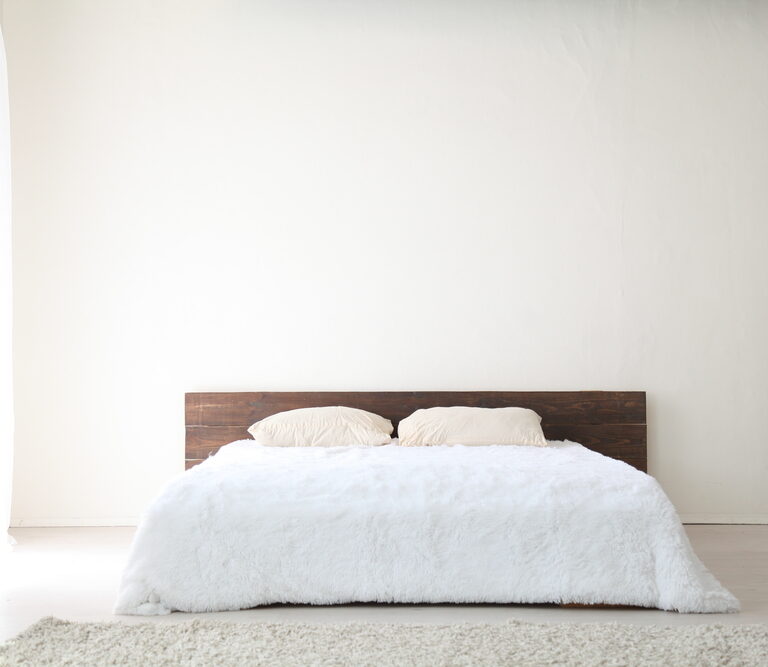 A minimalist bedroom with a low wooden bed frame, part of the Warehouse Advantage Library collection, features two beige pillows and a white fluffy comforter against a plain white wall. Soft natural light streams in, highlighting the cream rug on the floor.