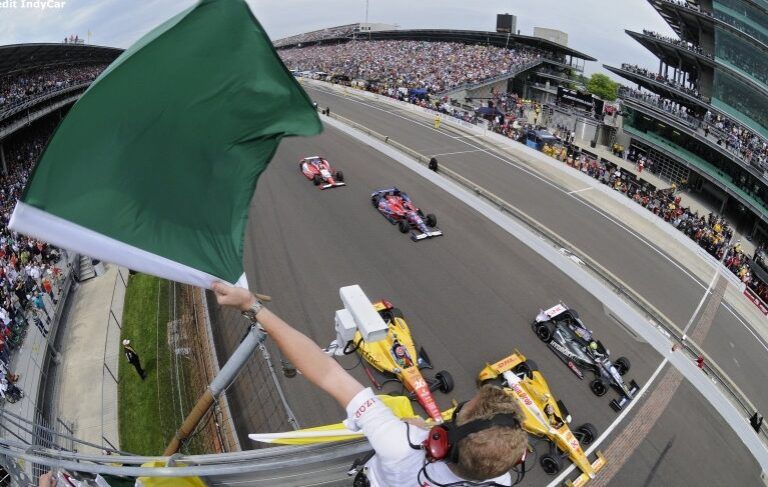 A race official waves a green flag to start an IndyCar race as several race cars speed past the starting line on a track, with a large crowd in the grandstands.