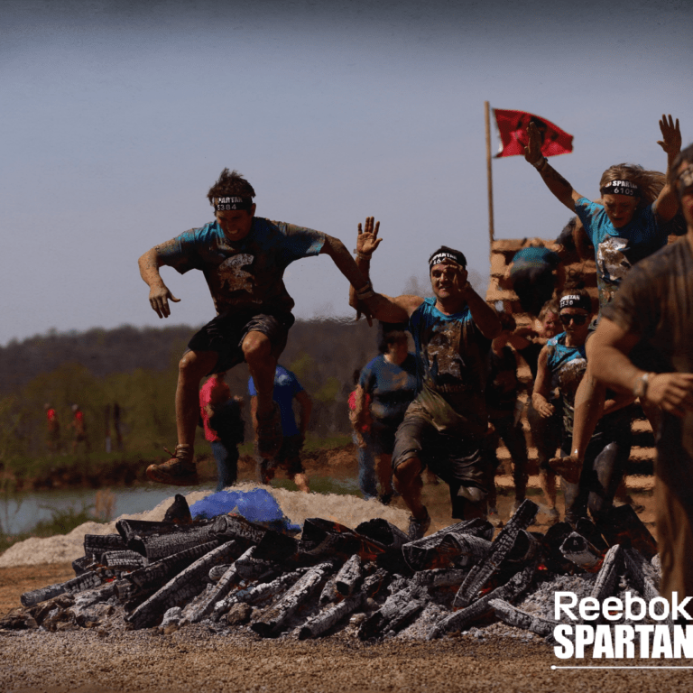 A group of athletes wearing headbands and muddy clothes jump over flaming logs during a Reebok Spartan Race obstacle course outdoors. The event logo is visible in the bottom right corner.