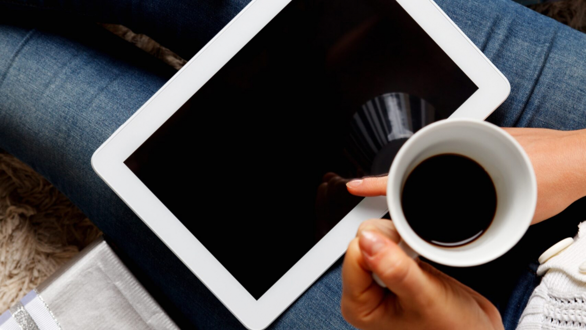 A person sitting with wrapped gifts holds a cup of coffee and a tablet, with a credit card nearby, suggesting online holiday shopping or gifting.