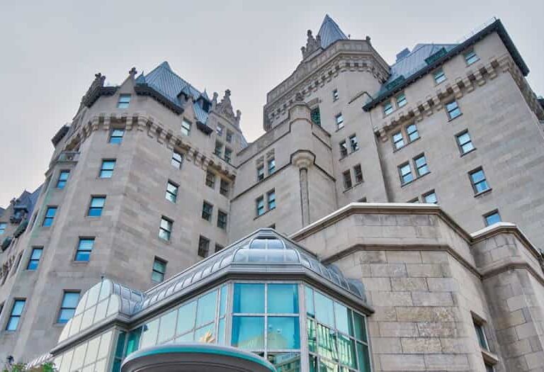 A view looking up at a large, historic stone building with turrets and steep blue-gray roofs, featuring many windows and a modern glass entrance in the foreground.