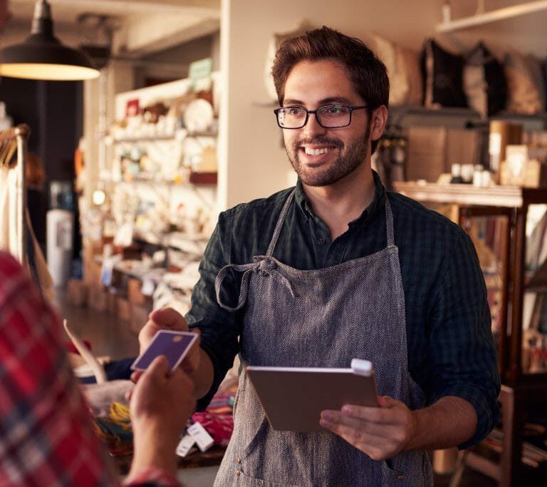 A smiling shop worker in an apron holds a tablet and processes a payment from a customer handing over a credit card in a cozy store environment. Shelves with various items are visible in the background.