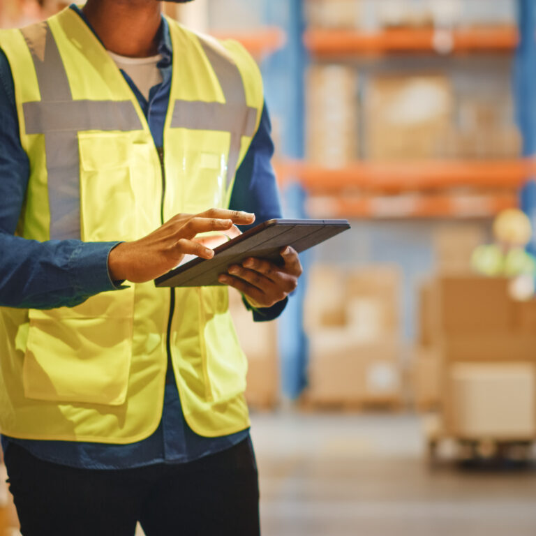 A person wearing a yellow safety vest uses a tablet in a warehouse filled with shelves and cardboard boxes.