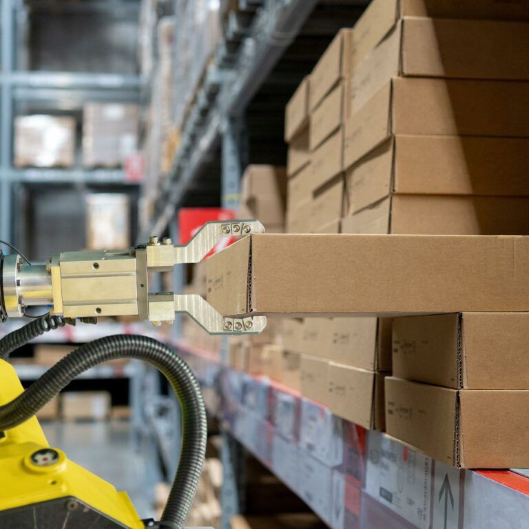 A robotic arm with a metal gripper lifts a cardboard box from a shelf in a dark warehouse filled with stacked boxes and shelves.