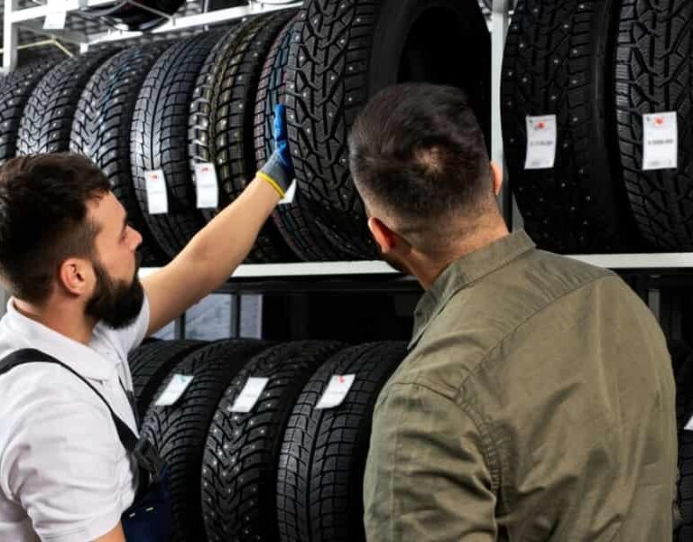 Two men examine and discuss tires displayed on shelves in a store; one man wearing a glove points at a tire while the other listens, with rows of tires and price tags visible.