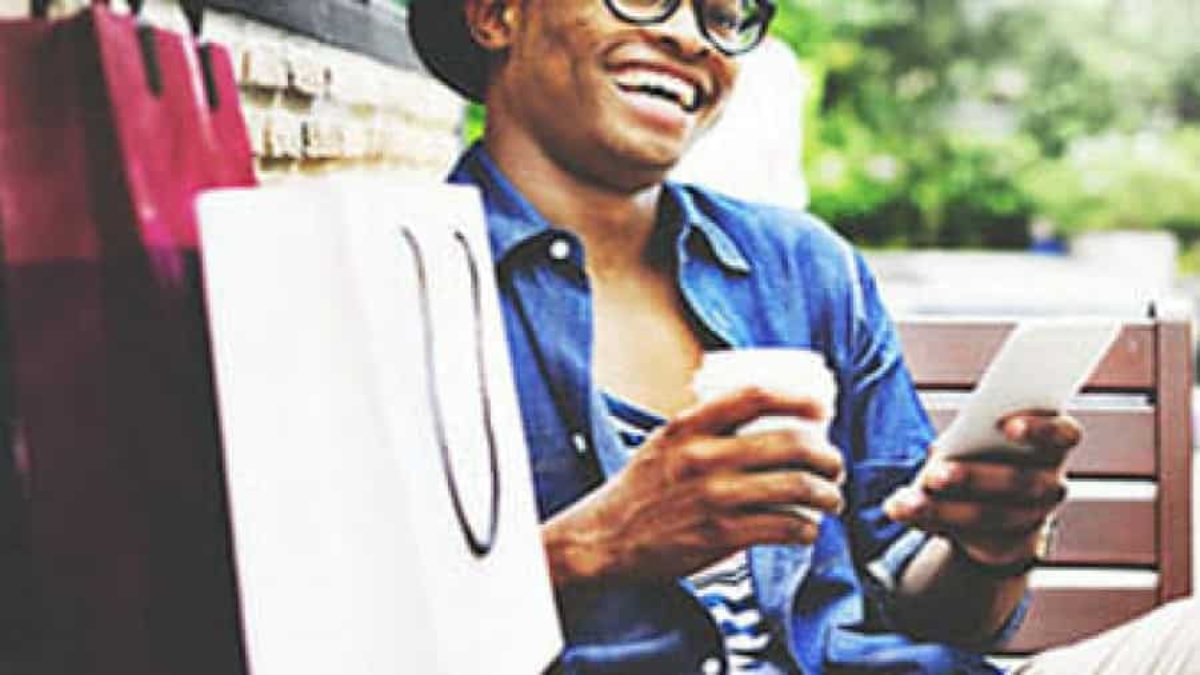 A smiling person wearing glasses and a hat sits on a bench holding a coffee cup and a smartphone, with shopping bags beside them. The person appears relaxed and happy, possibly enjoying a day out.