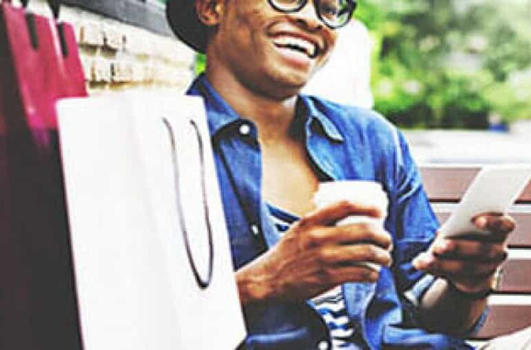 A smiling person wearing glasses and a hat sits on a bench holding a coffee cup and a smartphone, with shopping bags beside them. The person appears relaxed and happy, possibly enjoying a day out.