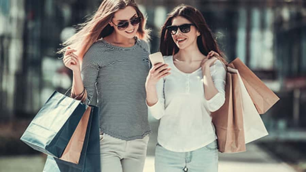 Two women wearing sunglasses are walking outside, smiling while looking at a phone. They are carrying several shopping bags and appear to be enjoying a day out shopping.