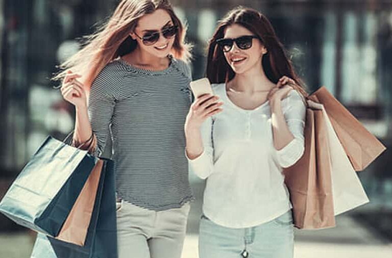 Two women wearing sunglasses are walking outside, smiling while looking at a phone. They are carrying several shopping bags and appear to be enjoying a day out shopping.