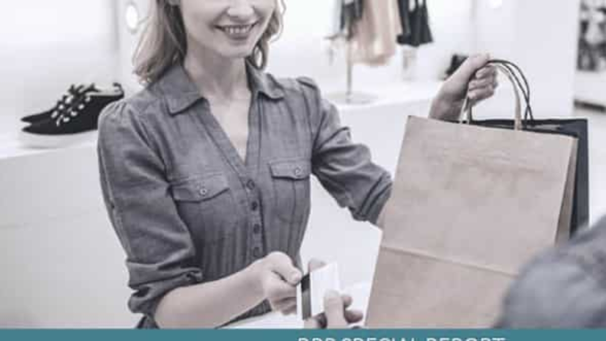 A smiling woman in a gray dress stands at a counter, handing a paper shopping bag to a customer while accepting a credit card for payment in a retail store.