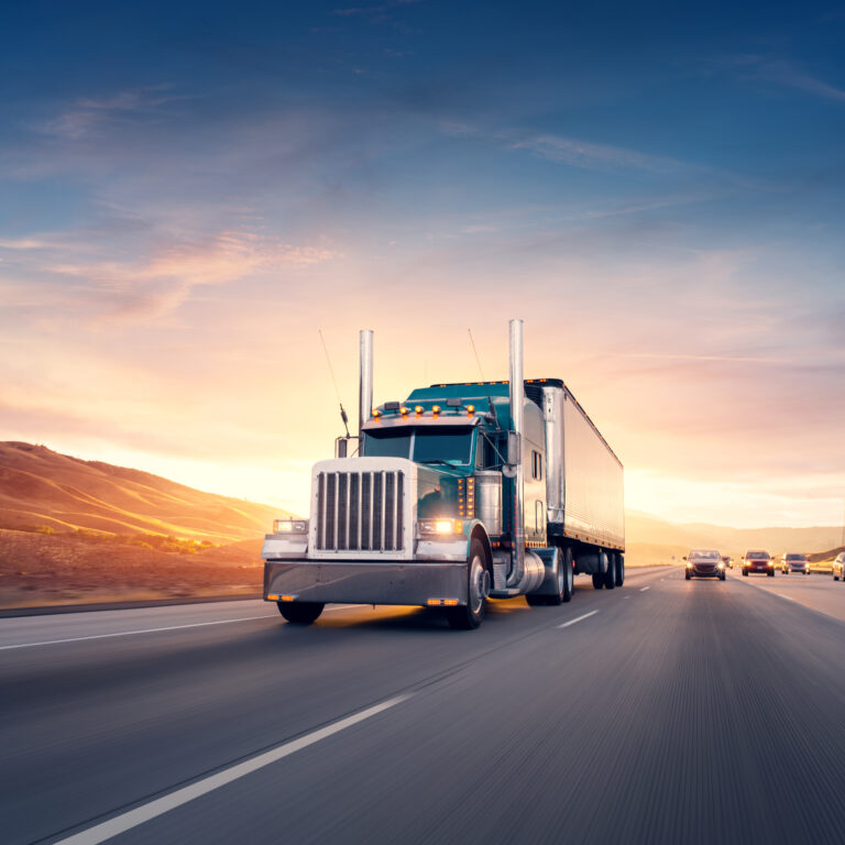 A large semi-truck drives down a wide highway at sunset, with hills in the background and several cars traveling in the same direction under a colorful sky.