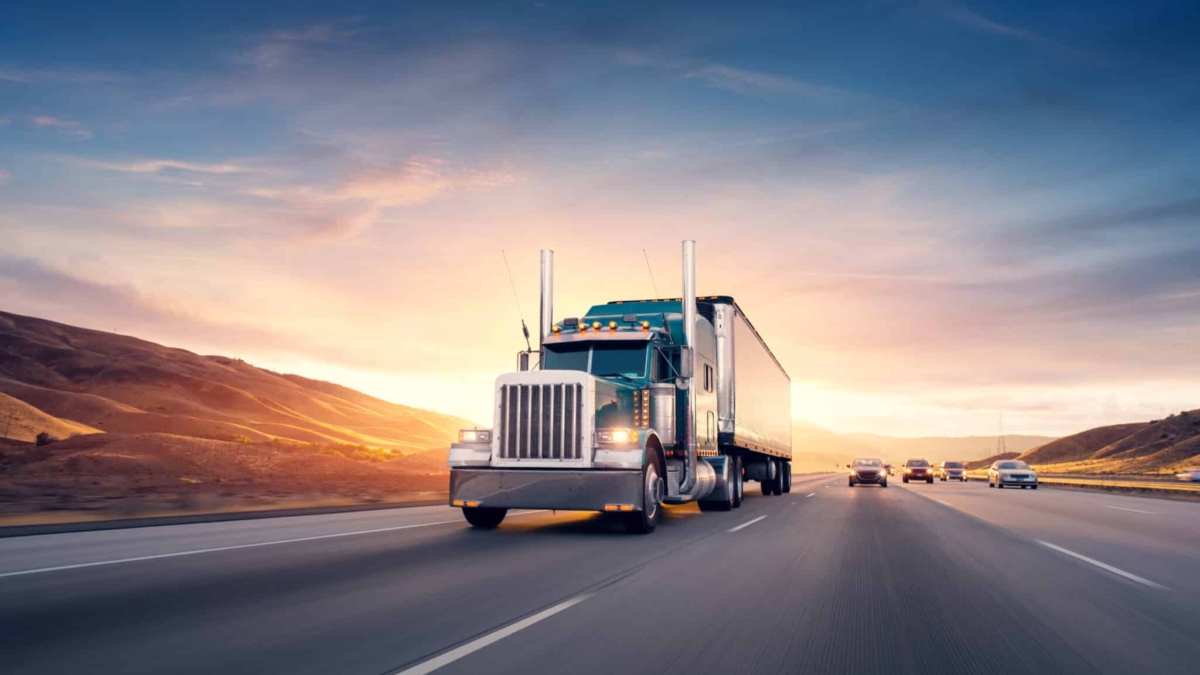 A large semi-truck drives down a wide highway at sunset, with hills in the background and several cars traveling in the same direction under a colorful sky.