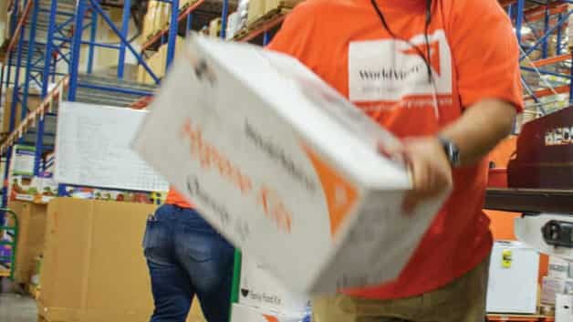 A person wearing an orange shirt carries a large white disaster kit box in a warehouse filled with shelves and supplies. Another person is working in the background.