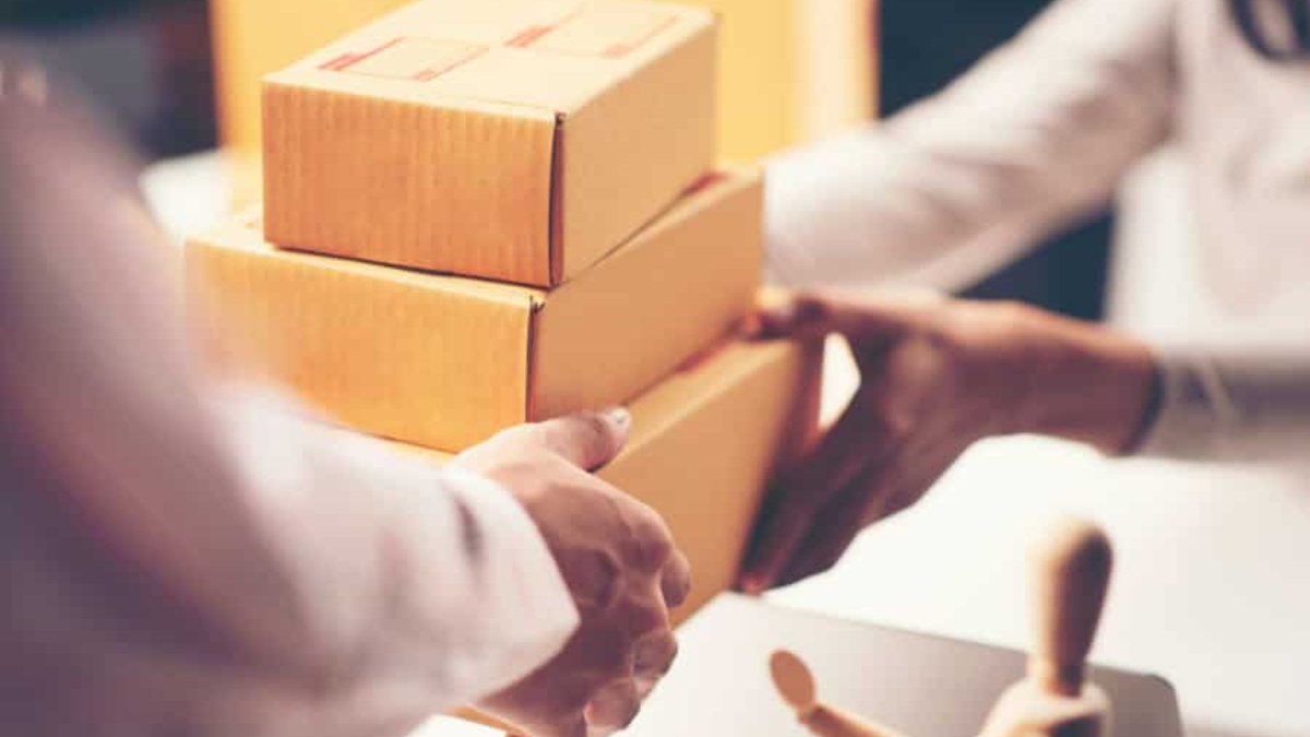 Two people exchange a stack of three small cardboard boxes, suggesting a delivery or package handoff. A wooden mannequin figure is visible on the table in the foreground.