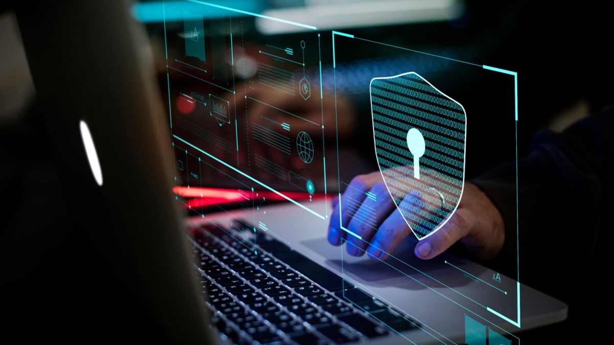 A person types on a laptop with a transparent digital shield and padlock overlay, symbolizing cybersecurity and data protection, highlighting the importance of avoiding the high cost of a security breach. Blurred computer screens and code fill the background.