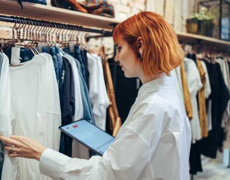 A person with short red hair wearing a white shirt uses a tablet while inspecting clothes hanging on racks in a clothing store.