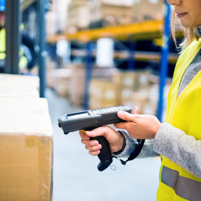 A worker in a yellow safety vest scans a cardboard box with a handheld barcode scanner in a warehouse. Shelves with more boxes and another worker in the background are visible.