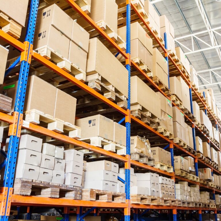 Rows of tall warehouse shelves filled with stacked cardboard boxes and packages, organized on wooden pallets in a brightly lit industrial storage facility.