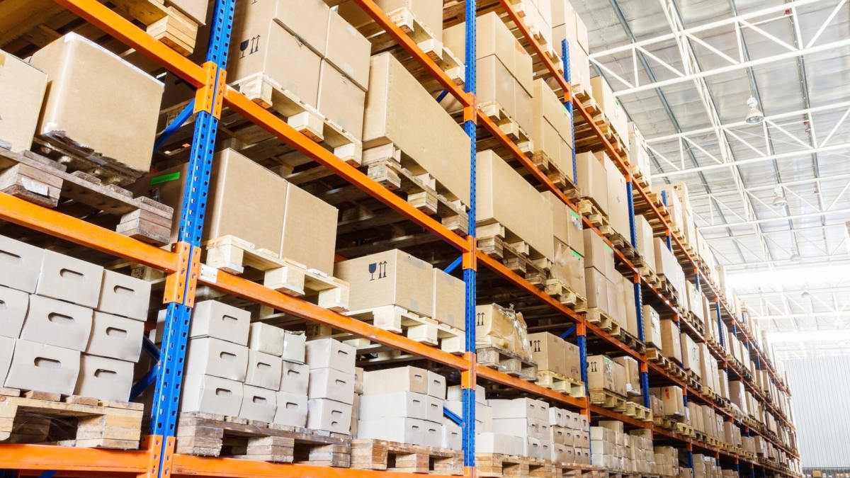 Rows of tall warehouse shelves filled with stacked cardboard boxes and packages, organized on wooden pallets in a brightly lit industrial storage facility.