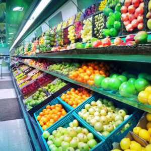 Shelves in a grocery store filled with a colorful variety of fresh fruits and vegetables, including apples, oranges, lemons, and peppers, neatly arranged in rows and baskets.