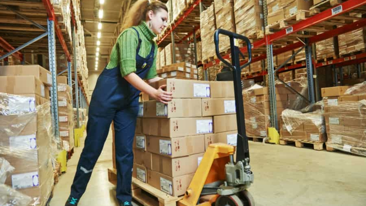 A woman in work overalls moves a pallet stacked with cardboard boxes using a pallet jack in a warehouse filled with shelves of boxed goods.