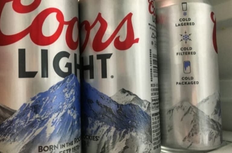 Three Coors Light beer cans with a mountain design are standing in a refrigerator. The cans are silver with red and blue accents, and the Coors Light logo is prominently visible on the front can.