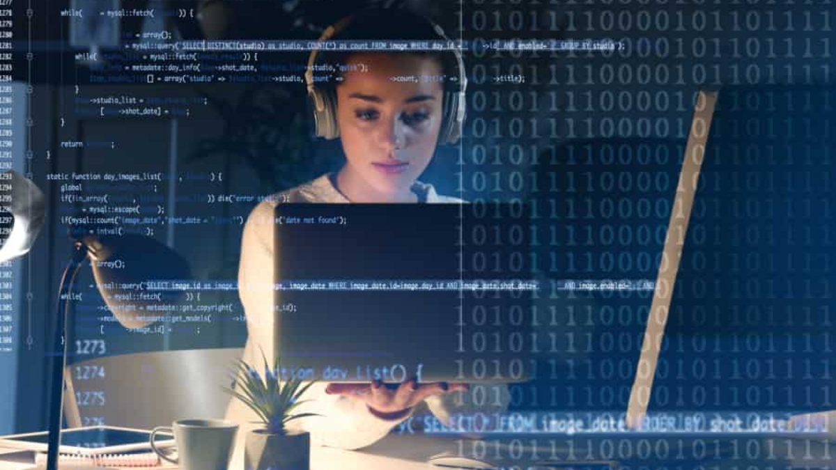 A woman wearing headphones works on a laptop at a desk, overlaid with blue computer code and binary numbers, suggesting programming or cybersecurity activity in a modern workspace.