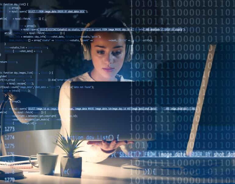 A woman wearing headphones works on a laptop at a desk, overlaid with blue computer code and binary numbers, suggesting programming or cybersecurity activity in a modern workspace.