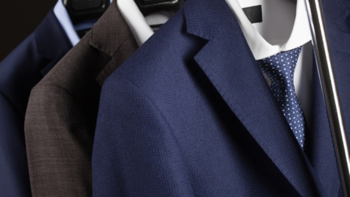 Three suits on hangers, including a blue suit with a white shirt and blue polka dot tie in front, and two darker suits behind it, all hanging on a clothing rack.