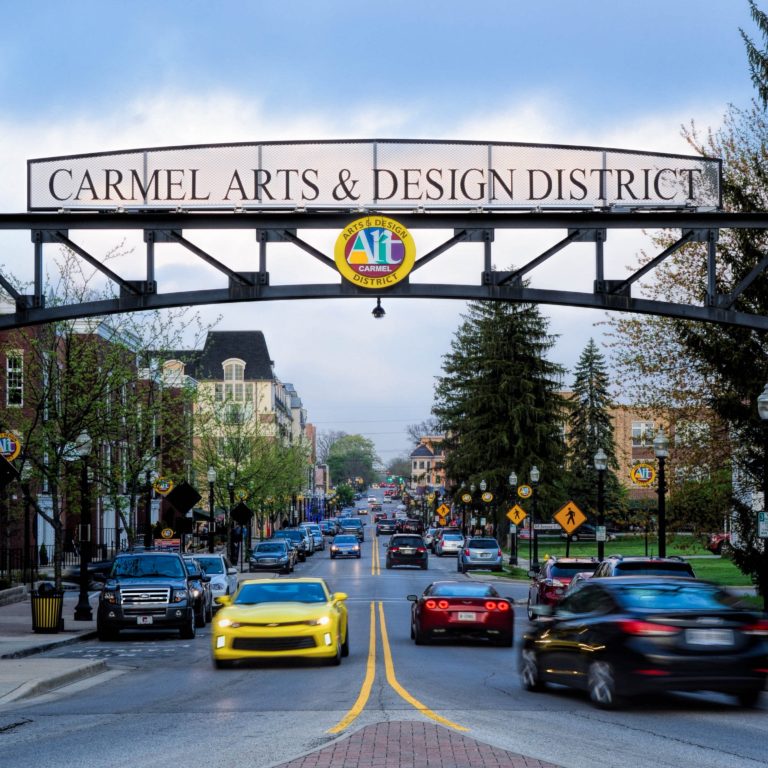 A street scene in Carmel Arts & Design District, Indiana, with a large archway sign overhead, cars driving on the road, trees, buildings, and people walking on the sidewalks. A yellow car is prominent in the foreground.